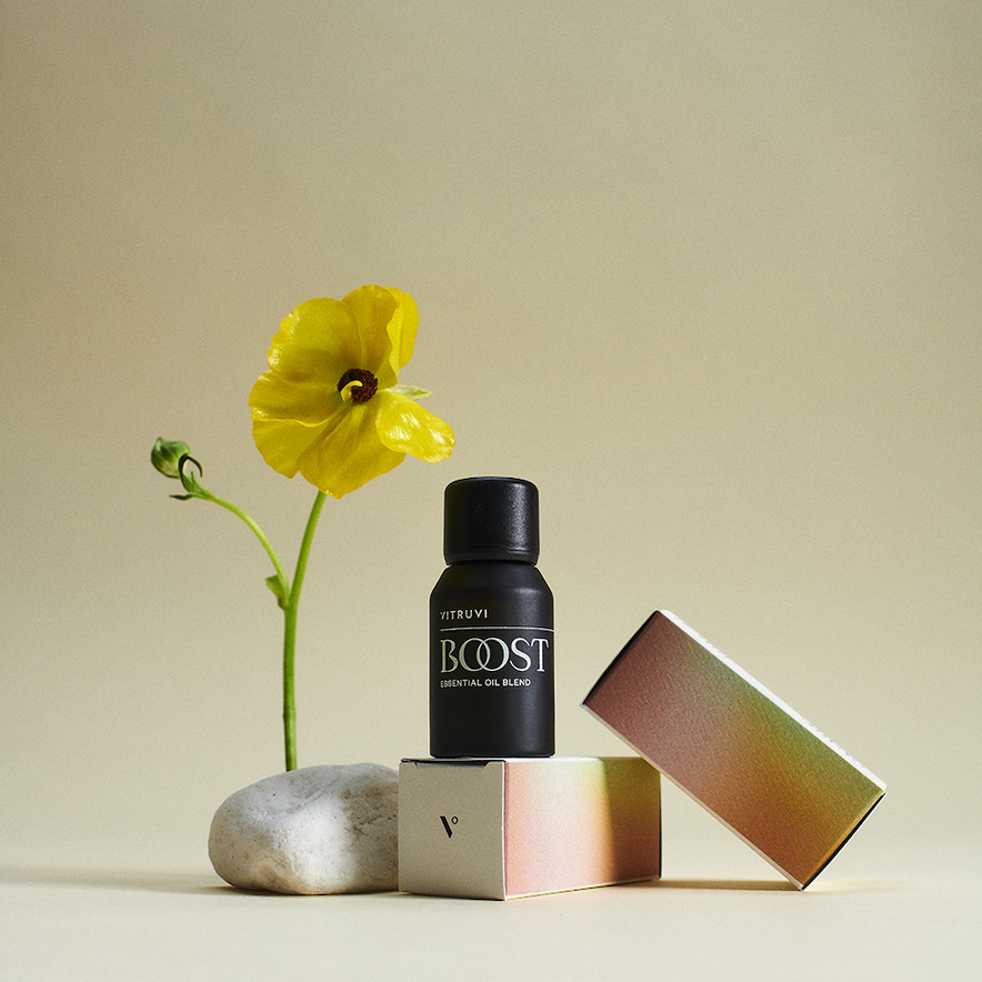The 'Boost' essential oil blends sits on top of a box. Another box, and a yellow flower frames the bottle. 