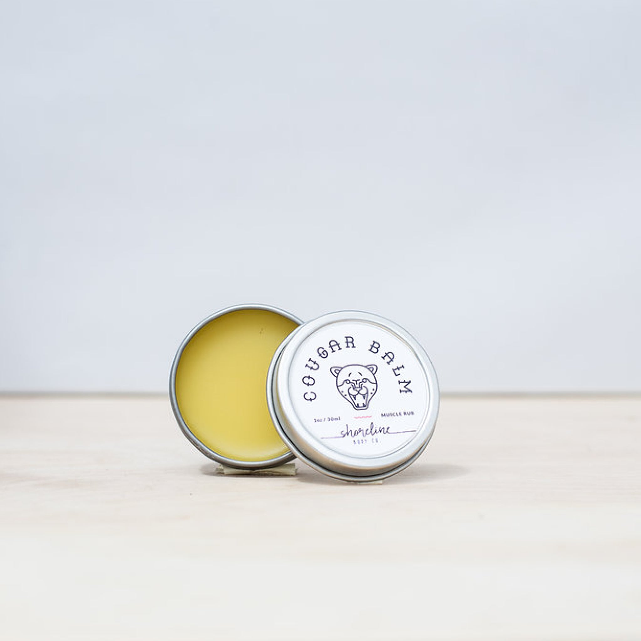 Cougar Balm, a soft pale yellow salve, is featured cap off with the lid leaning against it. 