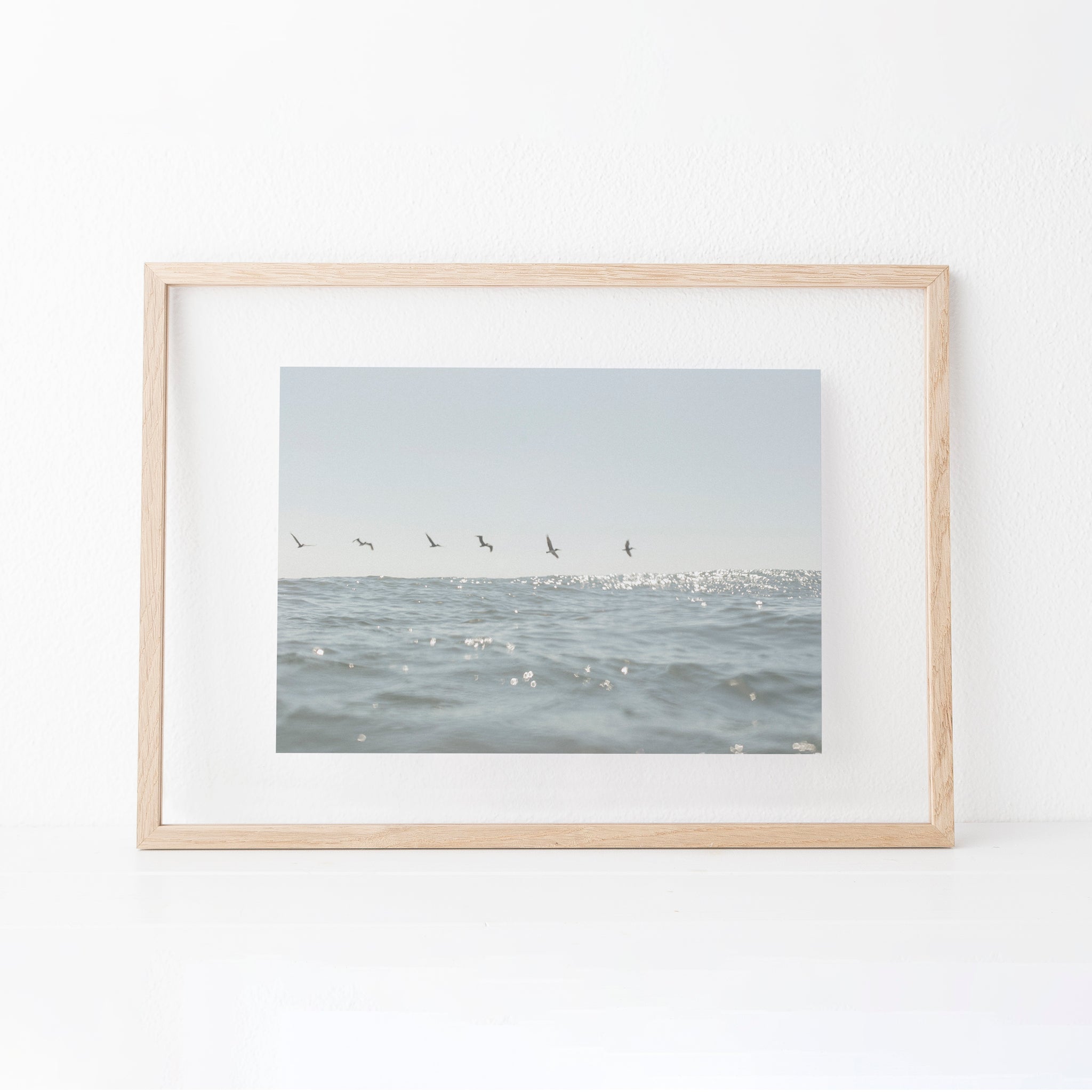 A photography print featuring shimmering light blue water and birds flying overhead. Photography by Cristina Gareau. 