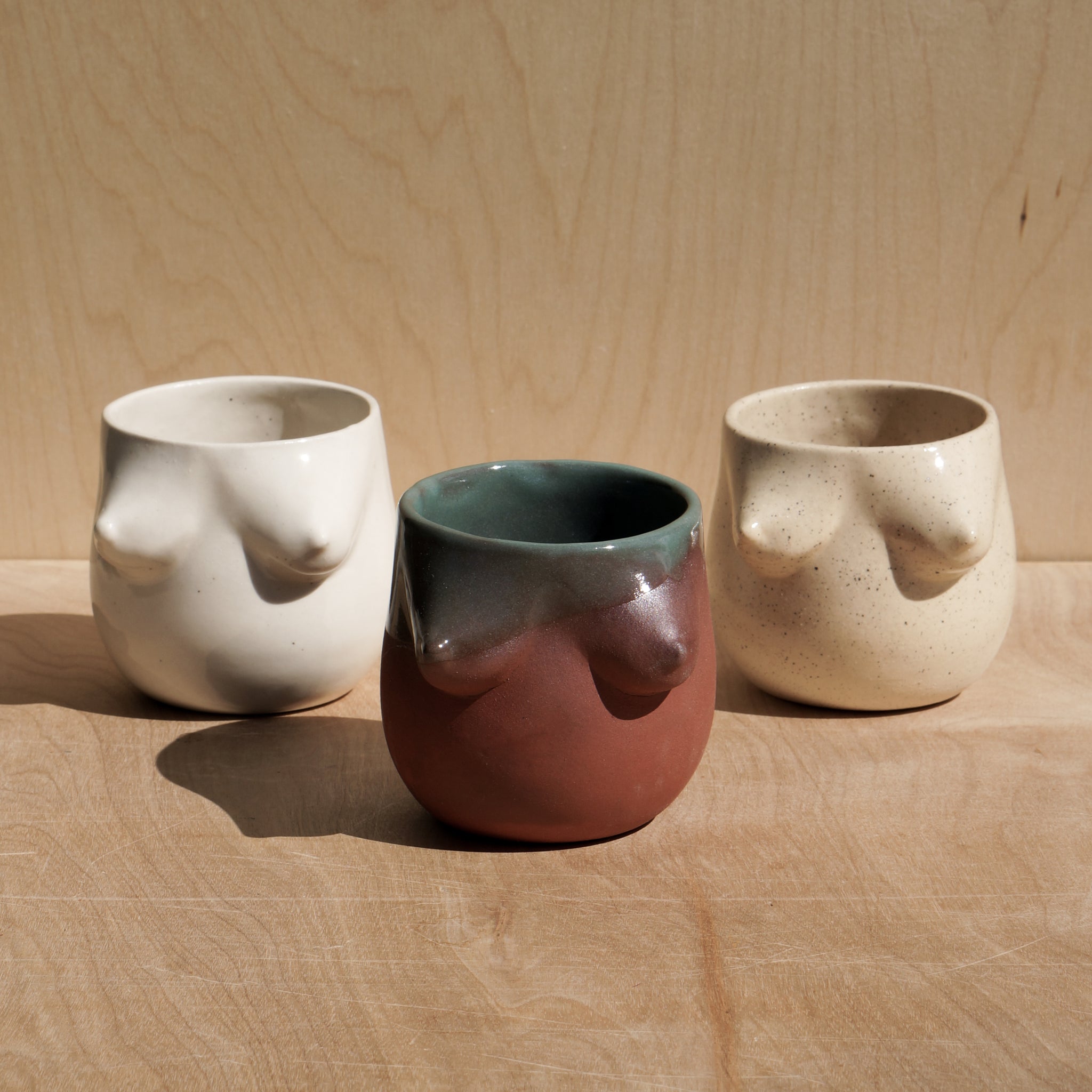 Ceramic tumblrs featuring the female form are pictured next to one another. From left,  a white ceramic tumbler, a rust red with a teal glass dripping down the side, and a speckled off white tumbler. 