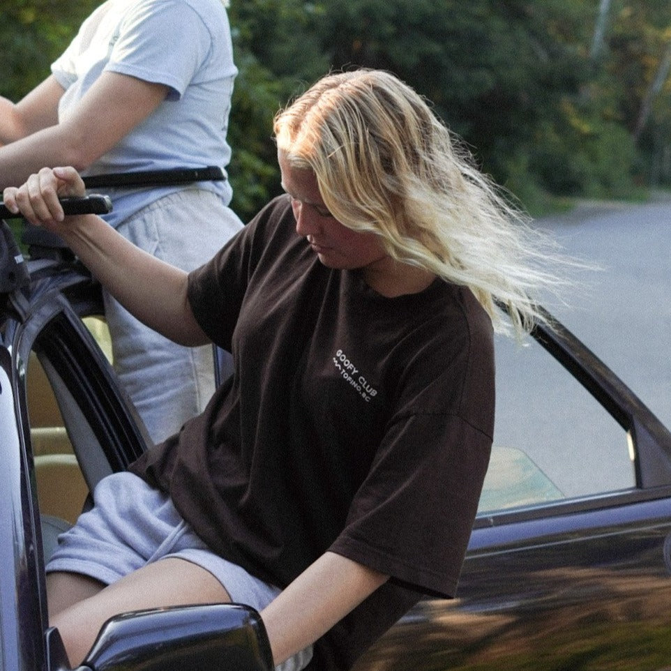 Blonde Model out car window wearing Goofy Apparel Dark Brown T with back graphic