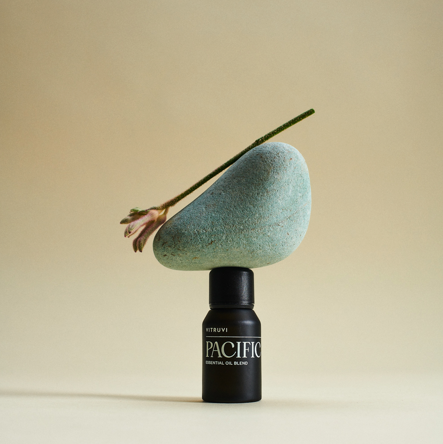 The 'Pacific' essential oil diffuser blend is pictured against an off-white background. On top of the bottle sits a smooth stone and a small clipping of a plant. 